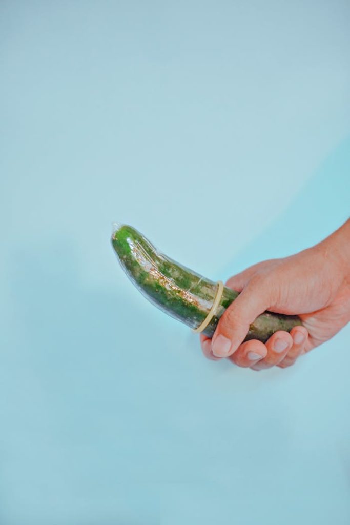 A cucumber wrapped with a condom held in a hand on a blue background, expressing safe sex concept.