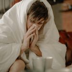 A young man wrapped in a blanket sneezes, indicating illness or cold symptoms.
