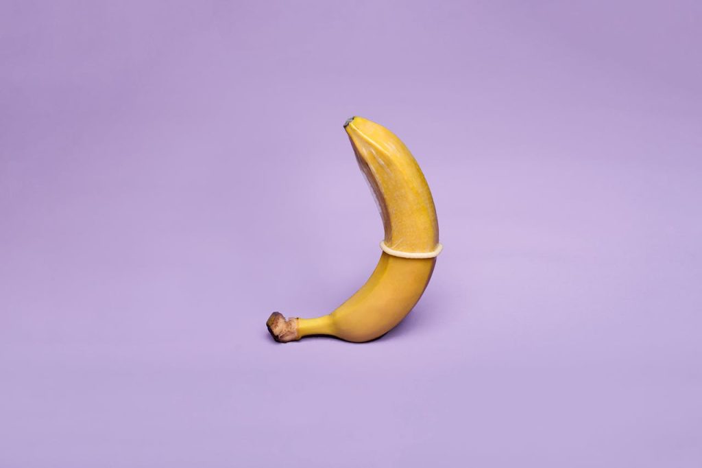 A banana wearing a condom symbolizes safe sex education against a pastel purple background.