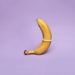 A banana wearing a condom symbolizes safe sex education against a pastel purple background.