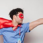 Male healthcare worker wearing a red cape and mask poses as a superhero. Studio shot.
