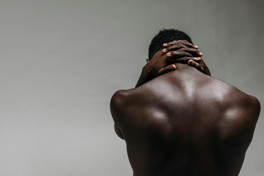 Back view of a brawny shirtless man with hands clasped on neck against a gray background.