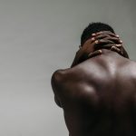 Back view of a brawny shirtless man with hands clasped on neck against a gray background.